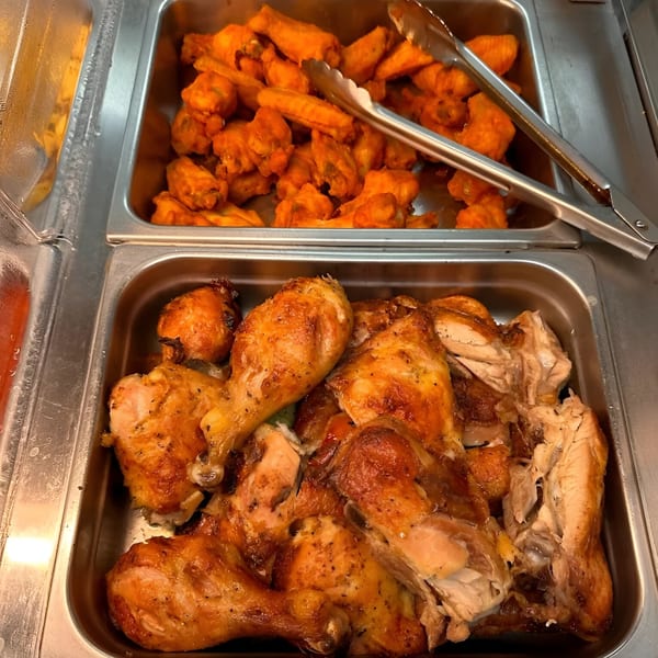 Two metal trays of food, one with golden-brown roasted chicken pieces and another with orange chicken wings.